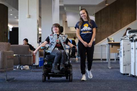 Two individuals moving through a library, one is a wheelchair user, the other is not.