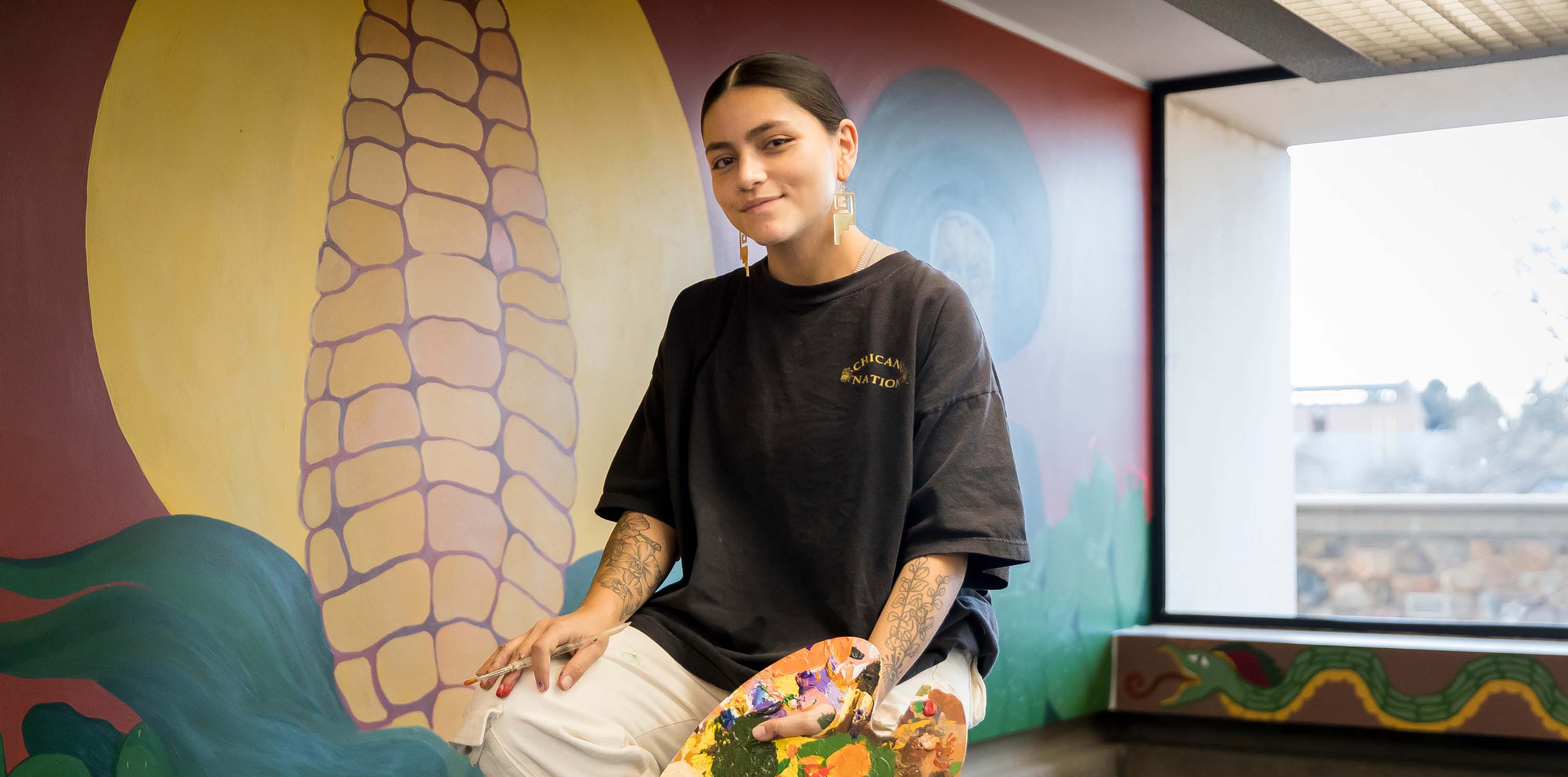 La alumna Brenda Vargas sentada en una escalera con un pincel frente a su mural