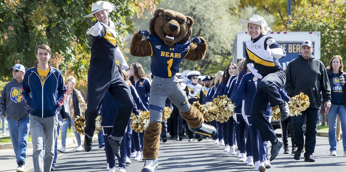 Klawz and two members from UNC's marching band jumping in the air while the band marches down a street.