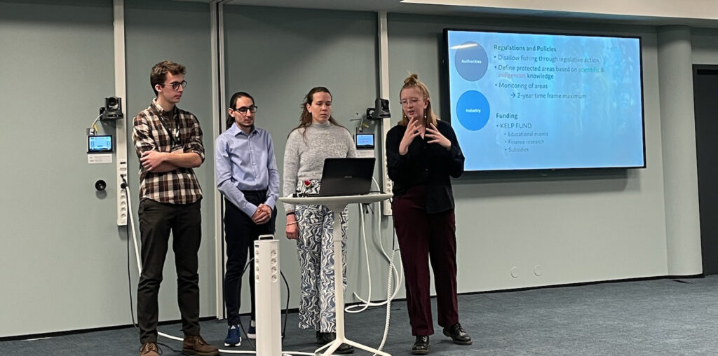 A group of four students standing in front of a presentation screen talking