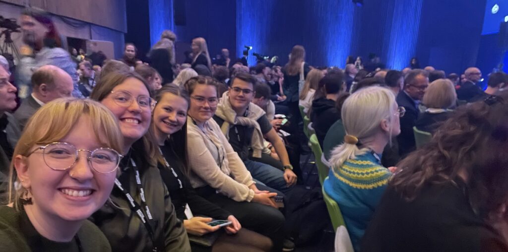 Dechant and Nichol sitting in an auditorium surrounded by other people