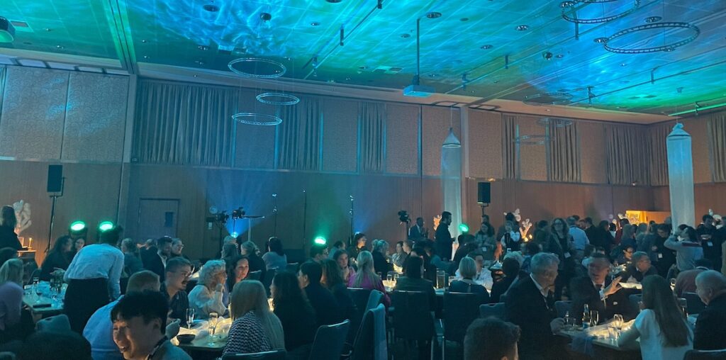 Interior of an events room lit with blue and green lights with people sitting at round tables.