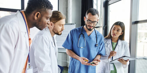 young male and female doctors in white lab coats taking notes and learning from older doctor in blue scrubs.