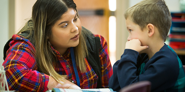 Hispanic/Latina teacher sitting beside young male student with a book