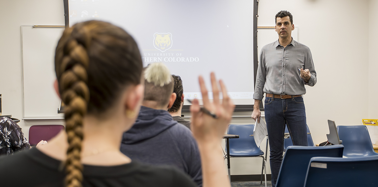 A young college student with their hand up in a classroom with the instructor standing at the front