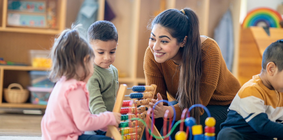Teacher sitting on the floor interacting with a group of preschool-age children.
