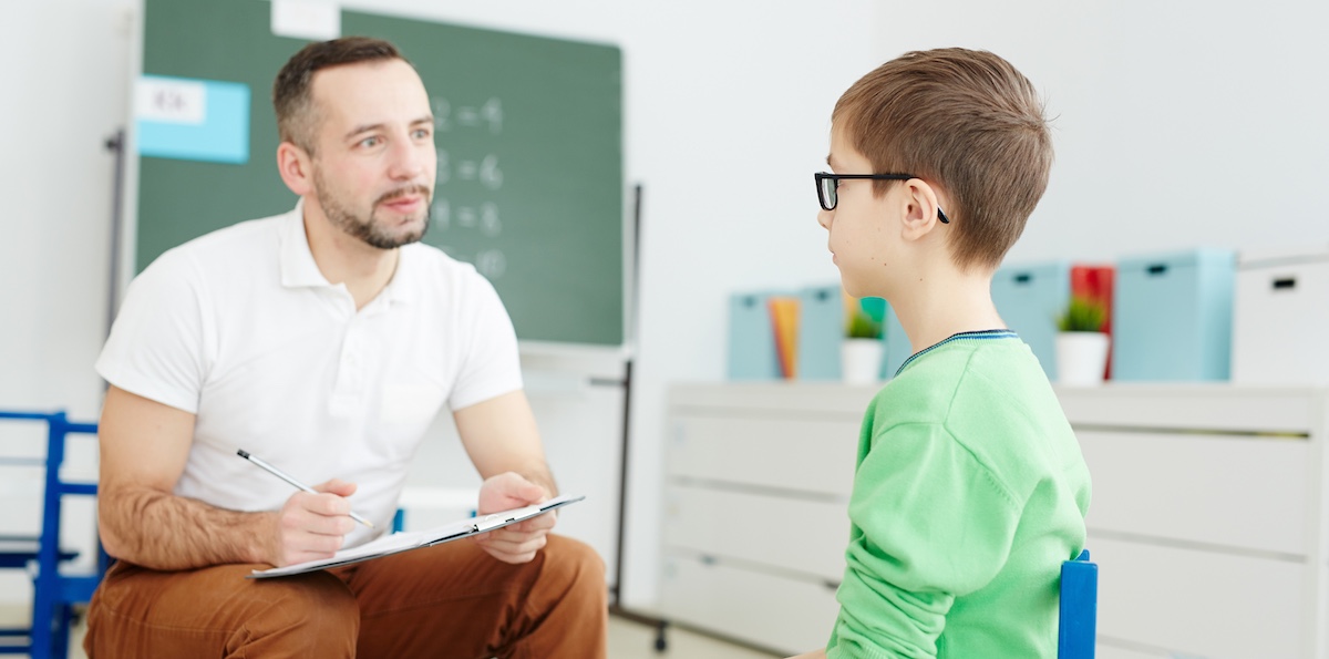 A male educator holding a clipboard sitting in a chair next to a young male student