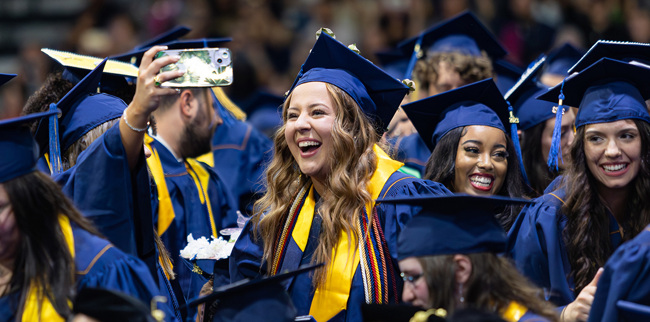 A group of UNC students standing and smiling and taking a selfie in their caps and gowns at graduation