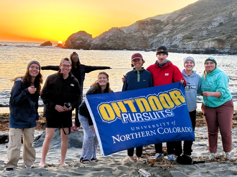 A group photo of students on the beach of Catalina Island in California for Spring Break with an Outdoor Pursuits flag at sunset.