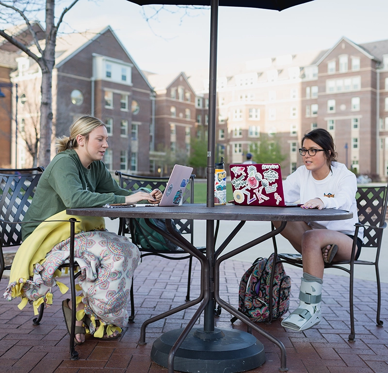 Two students sitting outside at a table working on laptops.