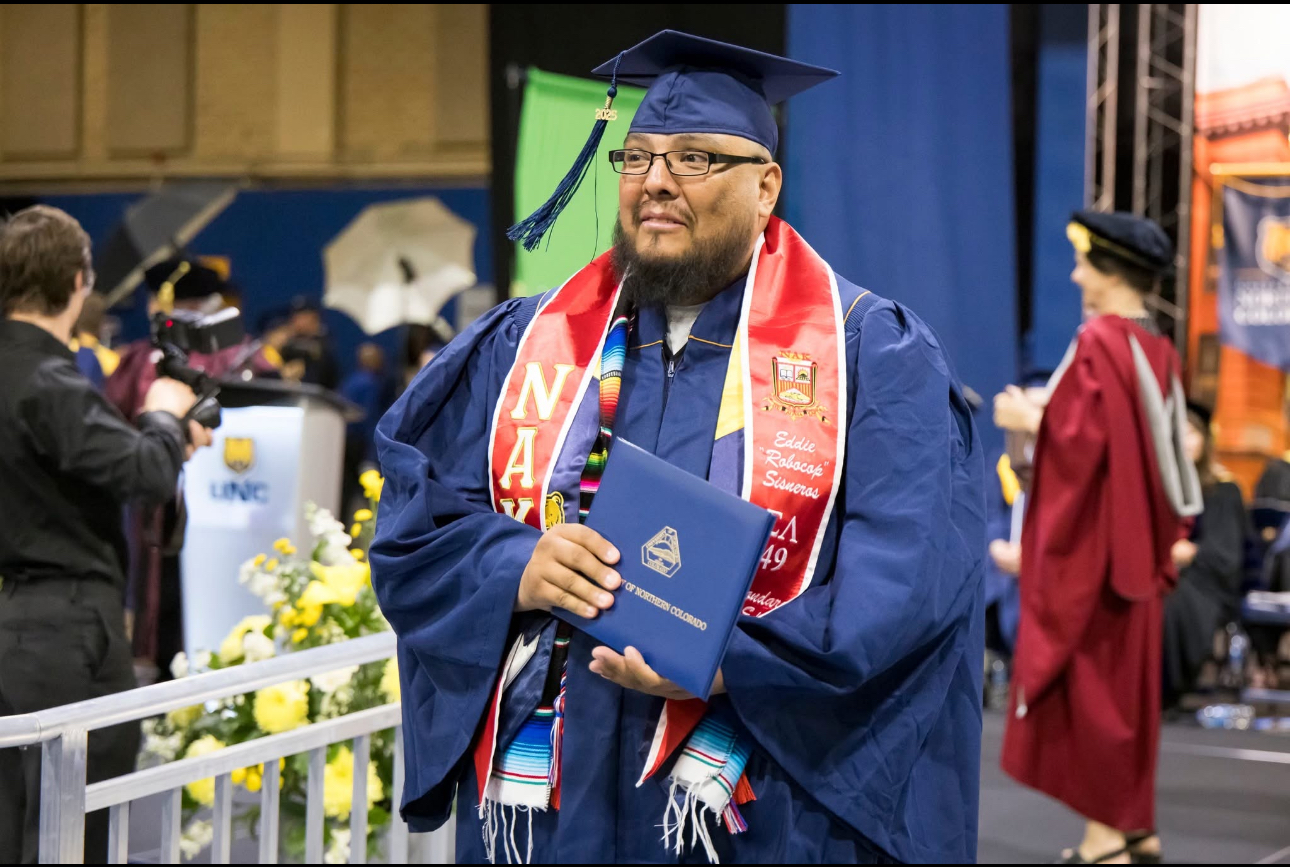 Graduate in cap and gown holds diploma during UNC ceremony, wearing a red “NAK” stole and colorful serape sash.