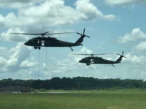 Two helicopters mid-air parallel to each other with rope dropped down from them.