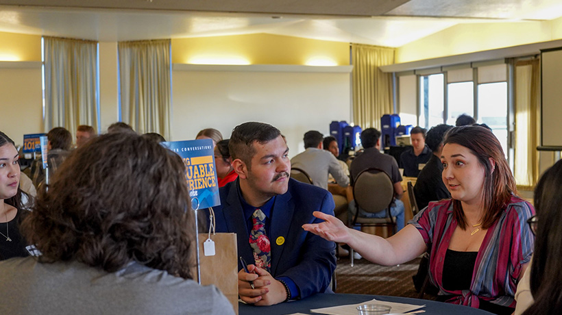 Two students engaged in a conversation during Career Conversations, with one student gesturing as they talk, while other participants work at tables in the background.