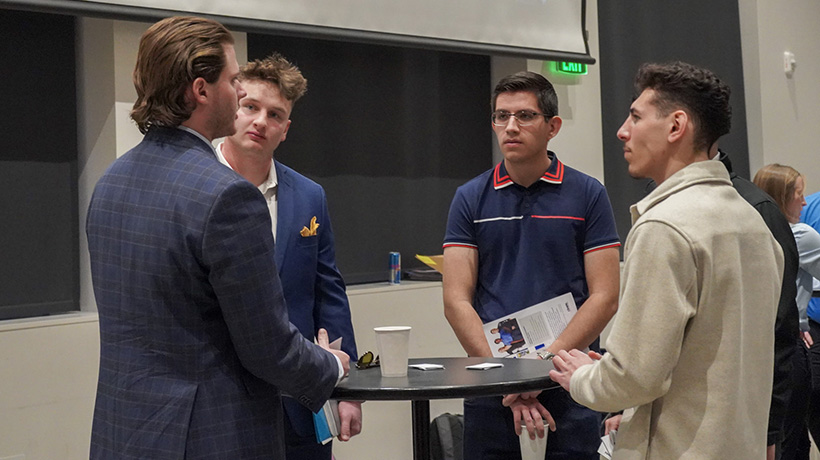 Four students, dressed in business casual attire, engaged in a conversation at Networking Night, standing around a table with cups and informational materials.