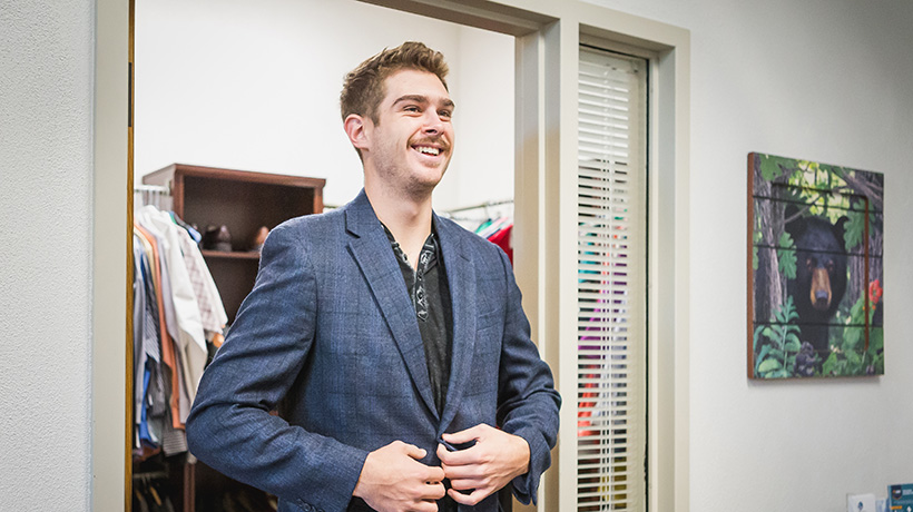 A young man smiling, wearing a blue suit coat, in the professional attire closet.