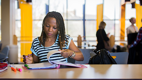 A young woman with dreadlocks studying and writing notes in a notebook at a table in a bright, modern campus setting.