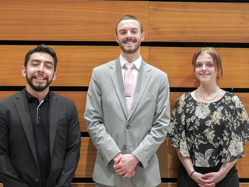 The top three students who won the 2024 Elevator Pitch Contest, dressed in professional clothing, standing against a wood background.