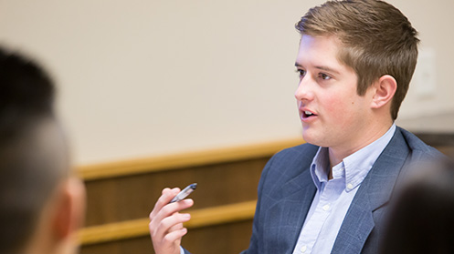 A young man in a suit jacket speaking and making a point during a discussion or interview in an indoor setting.