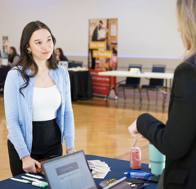 A young woman in a light blue cardigan and white top is listening attentively to another woman during a professional event, standing at a table with informational materials and a laptop.