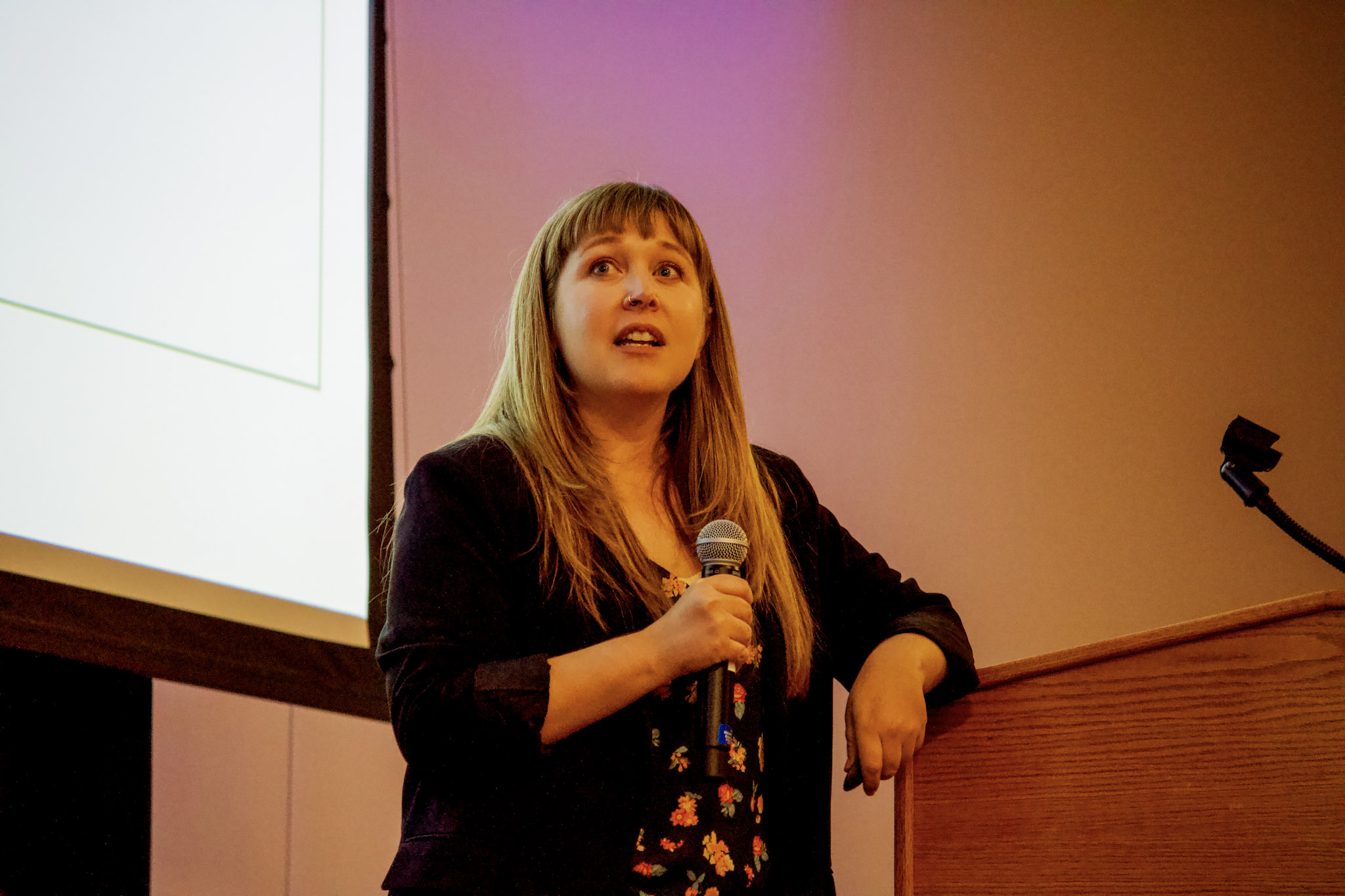A woman with long hair holds a microphone and appears to be speaking or presenting at a podium, with a projection screen behind her and a pinkish wall in the background. Regenerate Copy Good response Bad response