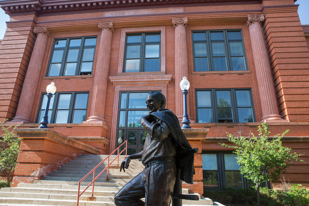 A bronze statue of a man leaning casually with his jacket slung over his shoulder is positioned in front of a red brick building with large windows and decorative columns. Two lampposts are on either side of a staircase leading up to the building's entrance.