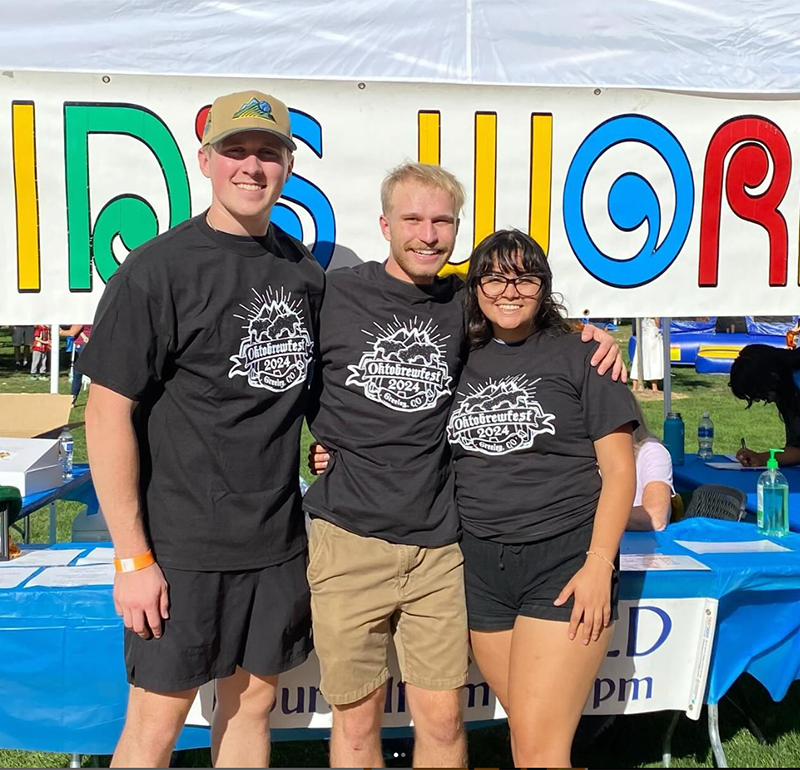 A group of students wearing matching black T-shirts, smiling, with a colorful background that resembles a fair setting.