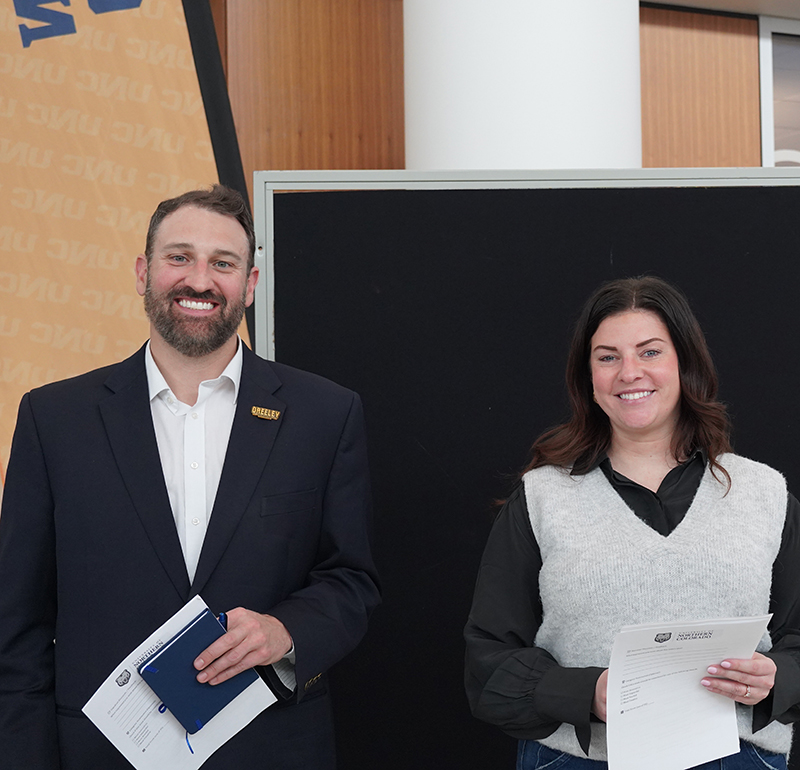 A man and woman dressed in professional attire, smiling while holding UNC-labeled papers.