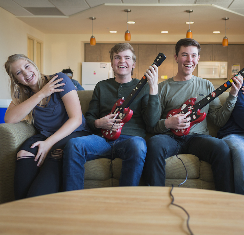 A group of three students sitting on a couch, laughing and enjoying a game of electric guitar.