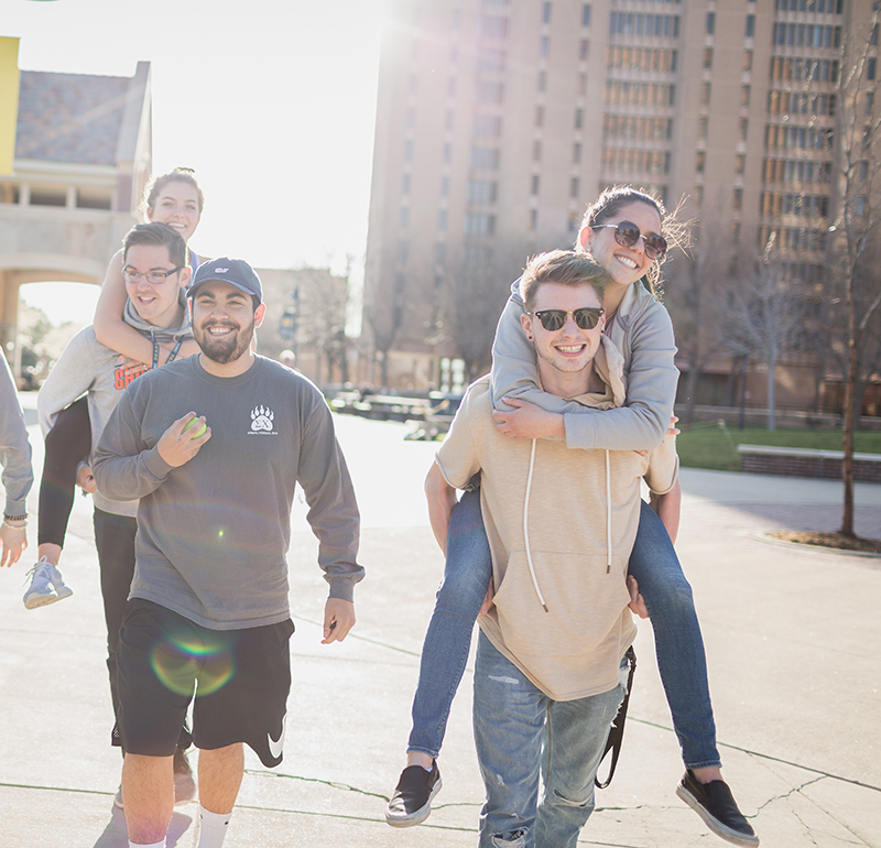 A group of young adults enjoying a sunny day outdoors in an urban park. One woman is riding on a man's back, both smiling and wearing sunglasses, with others walking and smiling in the background. Tall buildings are visible in the cityscape behind them.
