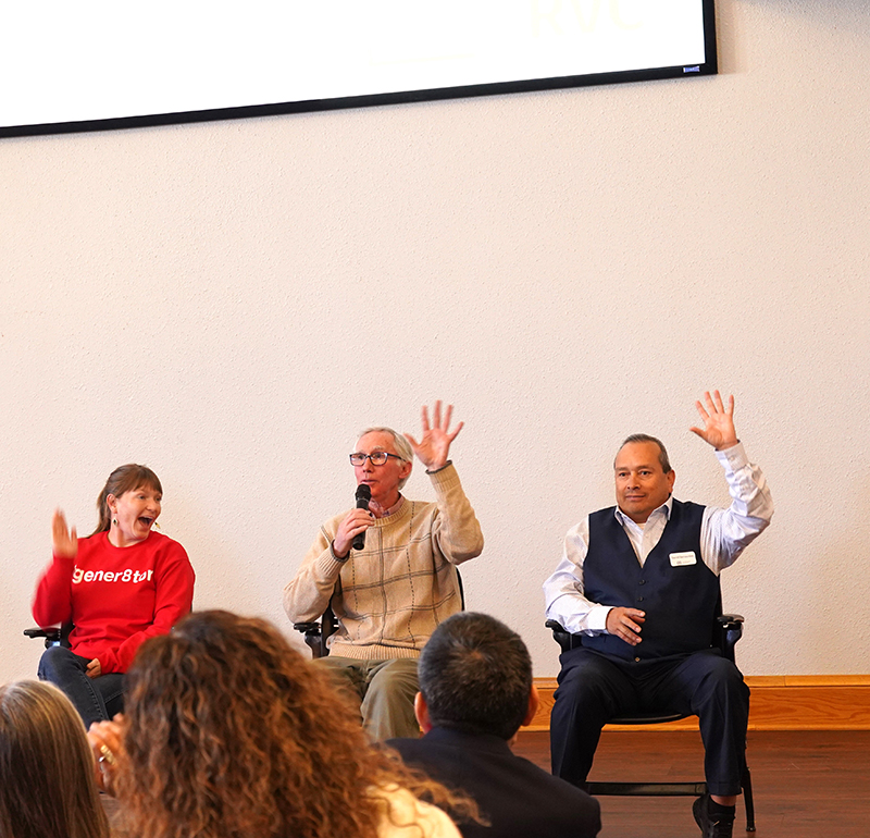 Three individuals seated on a stage, engaging with an audience. The person in the middle, holding a microphone, is speaking while the others raise their hands in response, with audience members visible in the foreground.