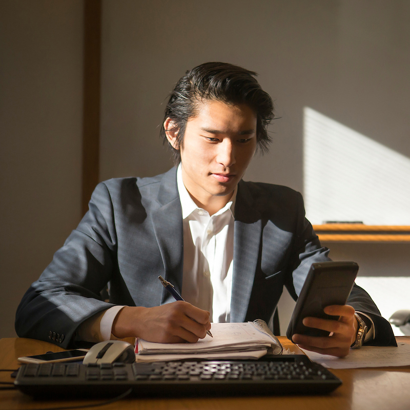 A young man with dark hair, dressed in a suit and white shirt, sitting at a desk, holding a phone in one hand and writing in a notebook with the other, illuminated by natural light. Concentrating on the task at hand — a UNC student in pursuit of knowledge.