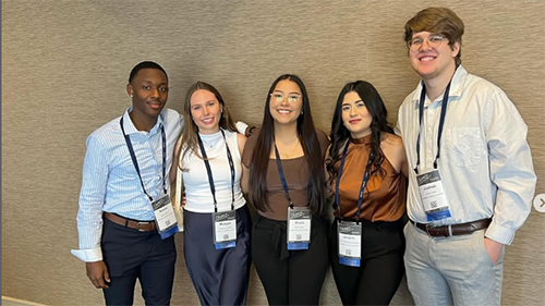 A group of students dressed in professional attire, smiling and wearing conference badges or name tags, suggesting they are attending a conference or formal event.