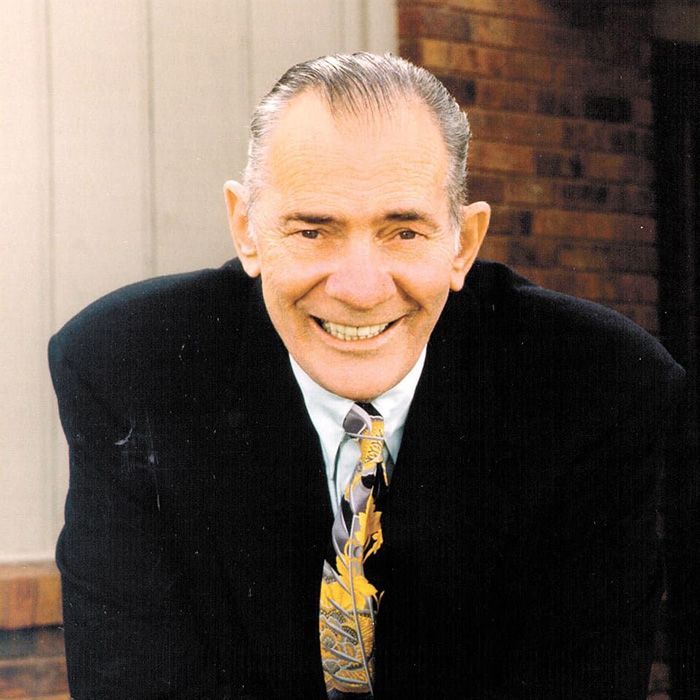 Kenneth Monfort smiling, dressed in a black suit and patterned tie, posing outdoors with a brick and wood-paneled building in the background.