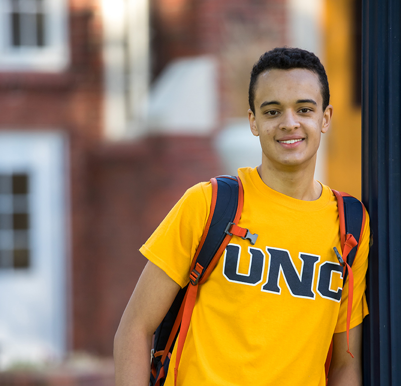 A male student wearing a yellow UNC shirt and carrying a red backpack, standing against a UNC background.