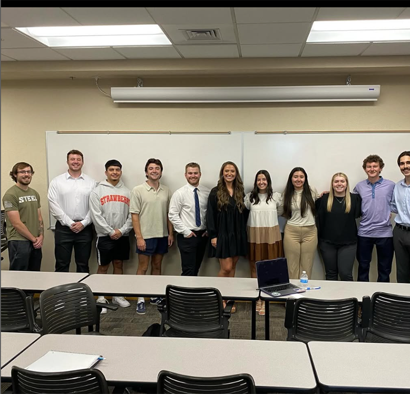 A group of students in a classroom standing behind a whiteboard, smiling, and dressed in professional attire.