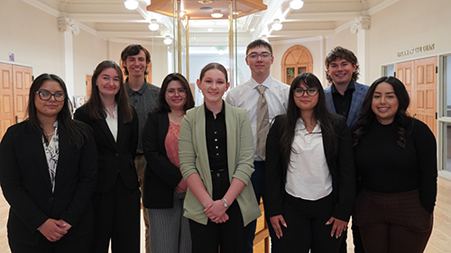 A group of ten young adults, dressed in business casual attire, standing together inside a building with elegant architecture. They are smiling at the camera, with some wearing glasses and others in blazer-style tops, suggesting a professional or academic setting.