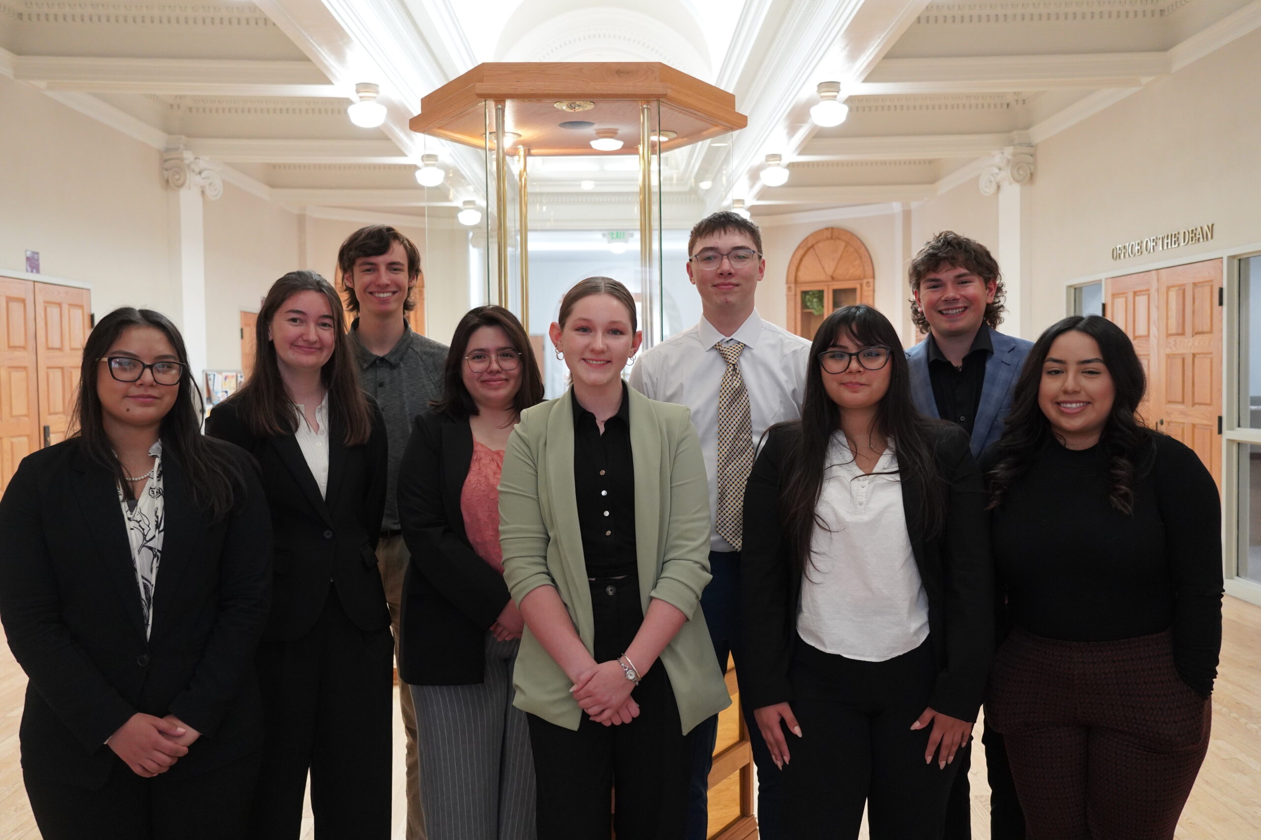A group of ten young adults, dressed in business casual attire, standing together inside a building with elegant architecture. They are smiling at the camera, with some wearing glasses and others in blazer-style tops, suggesting a professional or academic setting.