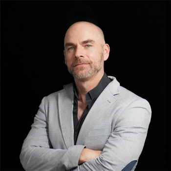 A man with a grey suit, crossing his arms and smiling. The background behind him is black.