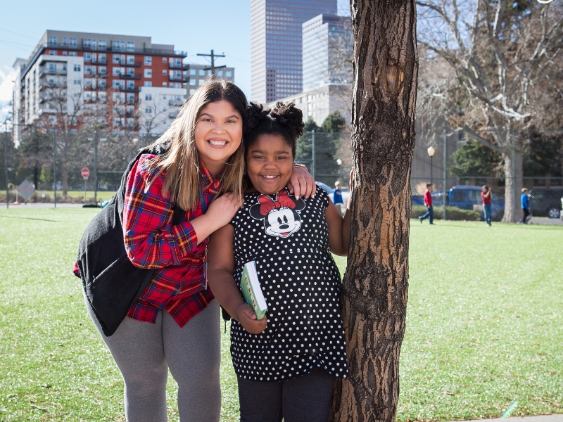 A center of Urban Education teacher is smiling at the camera with one of her students. They are standing next to a tree. The student is holding a book in her left hand smiling at the camera.