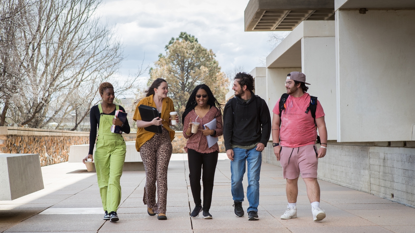 Five students are walking and talking across campus in front of the library.