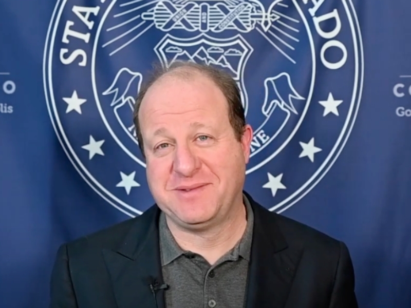 Colorado Governor Jared Polis is smiling at the camera with the State of Colorado flag behind him