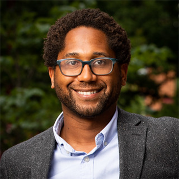 A man with curly hair, dressed in a grey suit jacket and blue shirt. Smiling with blurred trees in the background