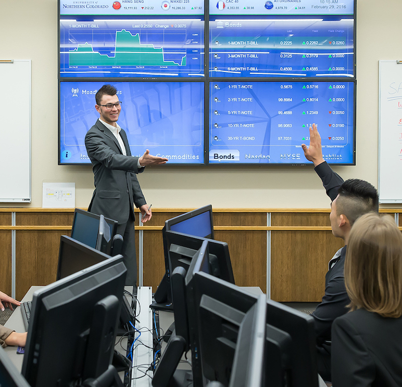 A group of students in a classroom setting with large screens displaying financial data. A student is raising a hand, and a presenter in a suit is gesturing towards the audience.
