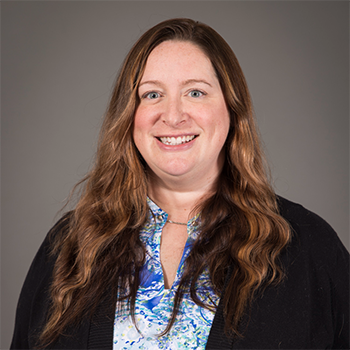 A woman with long red/brown hair, dressed in a colorful blue shirt. She is smiling and has a dark background behind her.