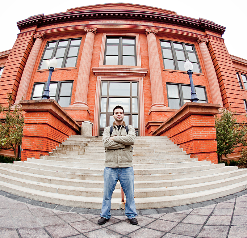 A male student smiling confidently with crossed arms, casually dressed, standing behind Kepner Hall—a brick building surrounded by trees and a light post.