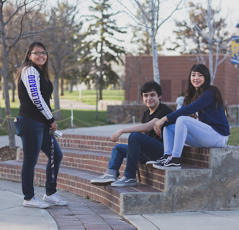 Three students, two sitting and one standing, dressed in casual clothing outside with a UNC flag in the background, hanging out and enjoying the day.