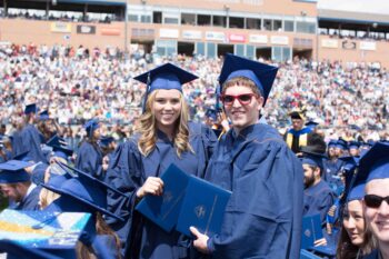 Two graduating UNC students pose with their degrees at an outdoor commencement ceremony.