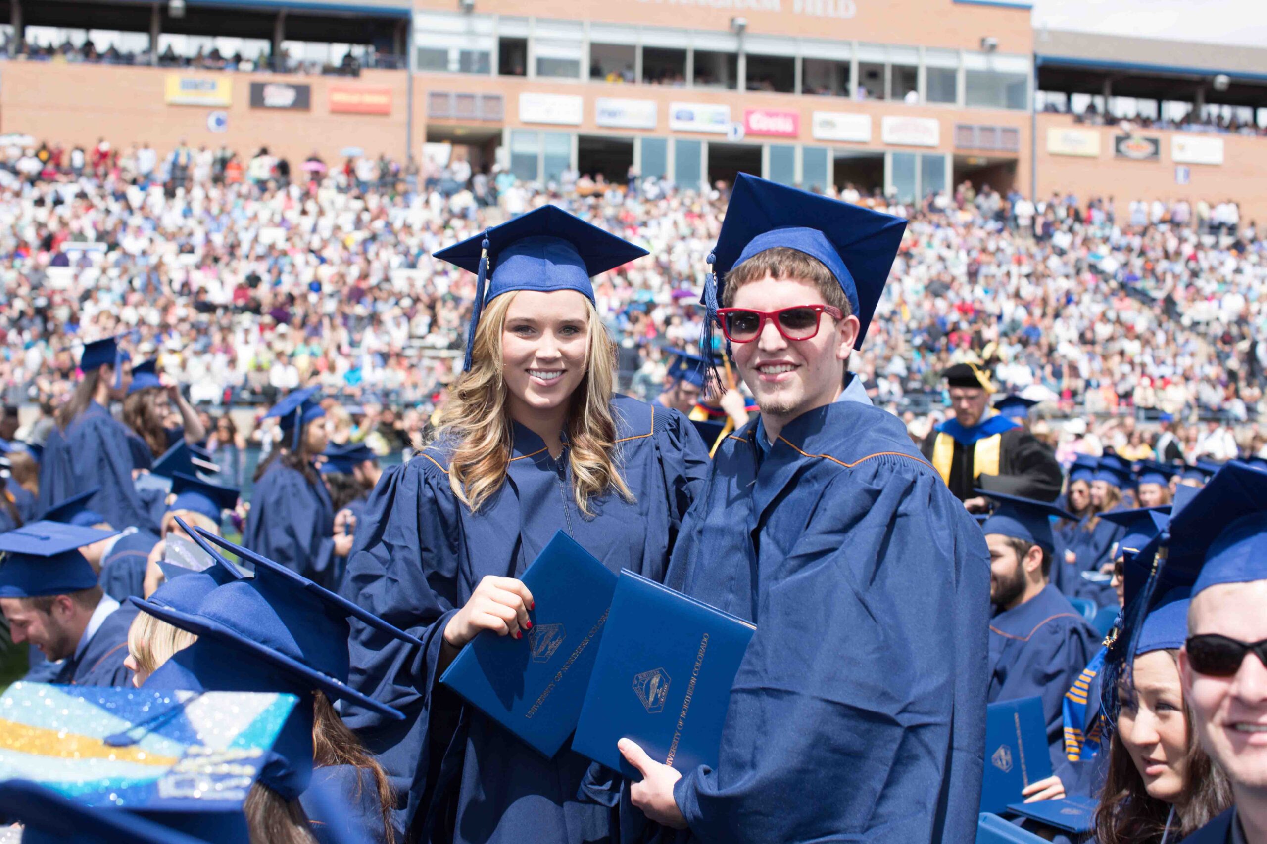 Two graduating UNC students pose with their degrees at an outdoor commencement ceremony.
