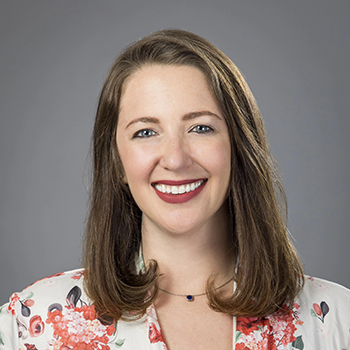 A women with medium length brown hair, a floral pink shirt and red lipstick, smiling.