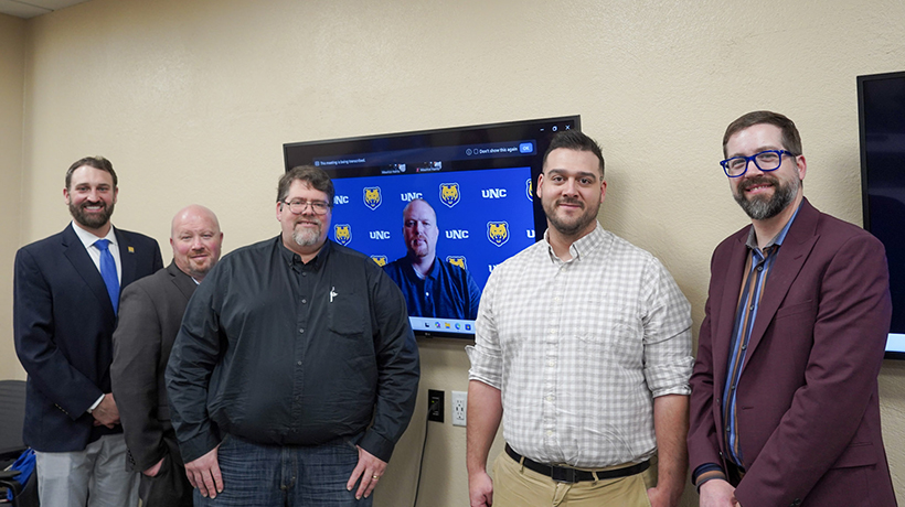 A group of five men standing in front of a screen displaying a video call participant, with the UNC logo in the background.